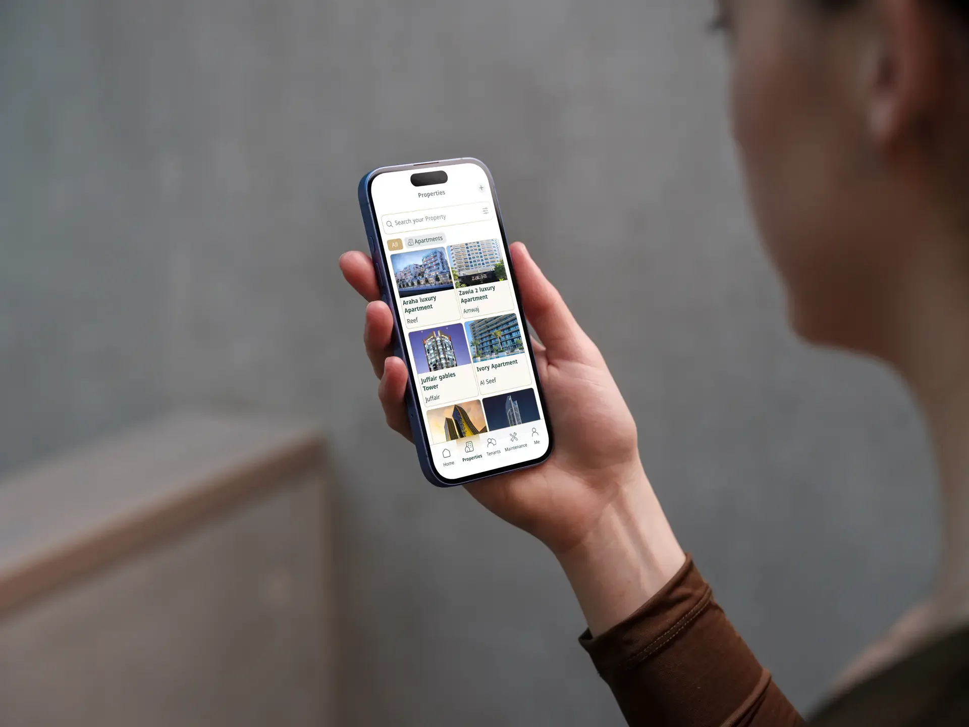 Person holding a phone showing Mullak+ property listings, soft-focus background